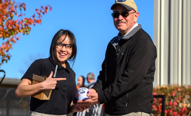 Portage School of Leaders High School junior Rebecca Anderson, wearing a black short-sleeved shirt and glasses, stands smiling beside NASA Glenn’s Daniel Sutliff, an acoustic engineer, on the campus of the University of Notre Dame. Sutliff is wearing a NASA ballcap and black jacket. He and Anderson are holding an autographed NASA mug, which Sutliff signed with a black ink permanent magic marker. Anderson is holding the black marker and a small cardboard box.