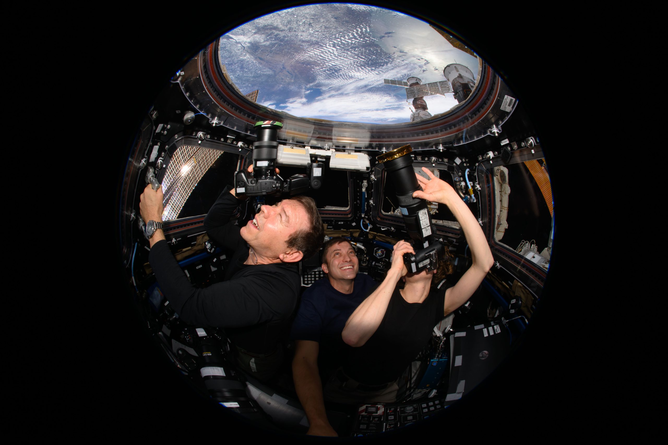Three astronauts, all wearing black, are facing the camera and looking out a cupola window, where Earth is visible above them. Barratt, on the left, holds a handrail with his left hand and points a camera toward Earth with his right hand, squinting his left eye. Dominick, center, smiles with eyes toward the window, and O’Hara, on the right, uses both hands to point a camera with a large lens at Earth. The camera obscures her face.