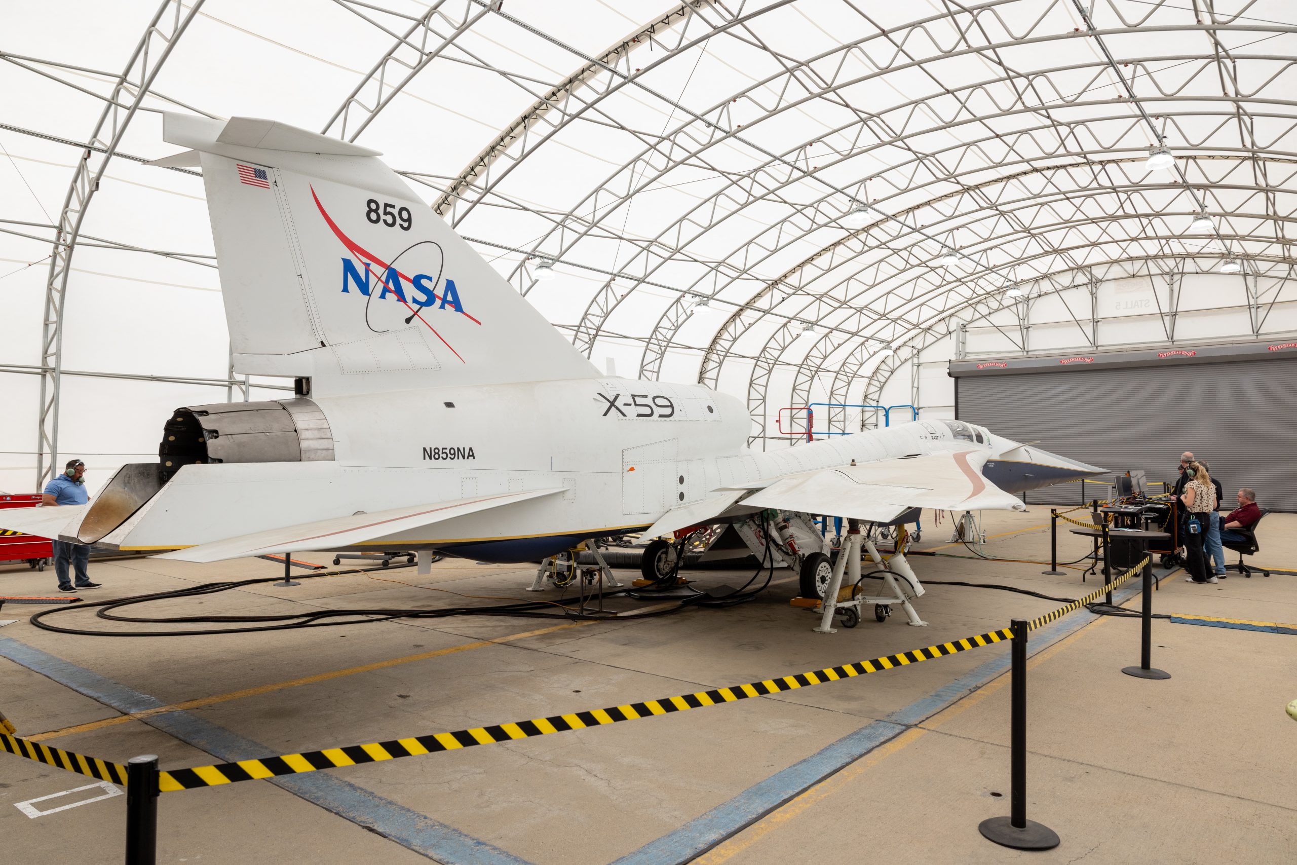 A white plane sits in a hangar that has a curved ceiling. The plane has markings on it including a NASA logo and "X-59." Several people are inside the hangar as well.