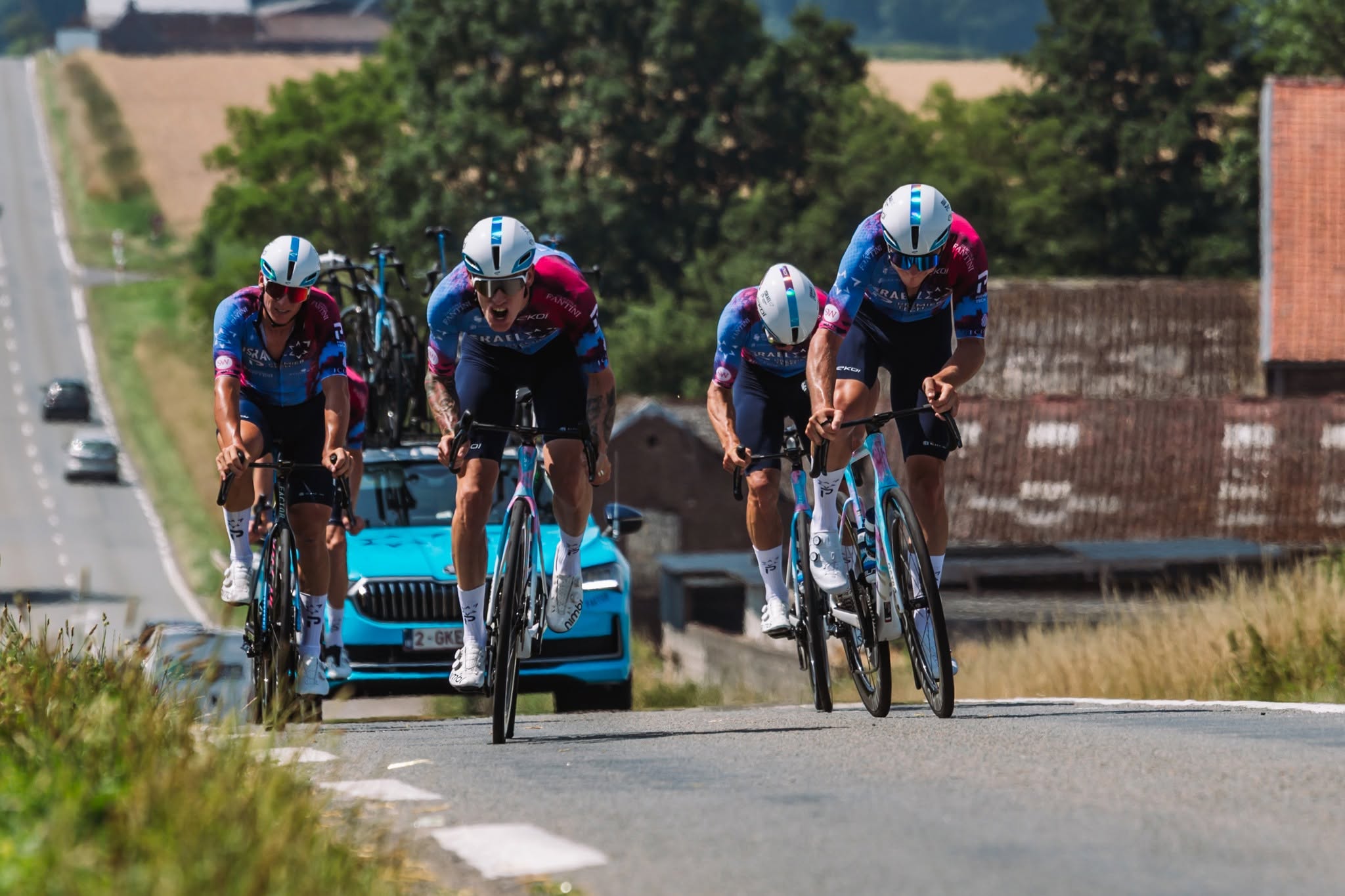 A team of racing cyclists clad in racing gear riding up a hill, a blue car driving behind them