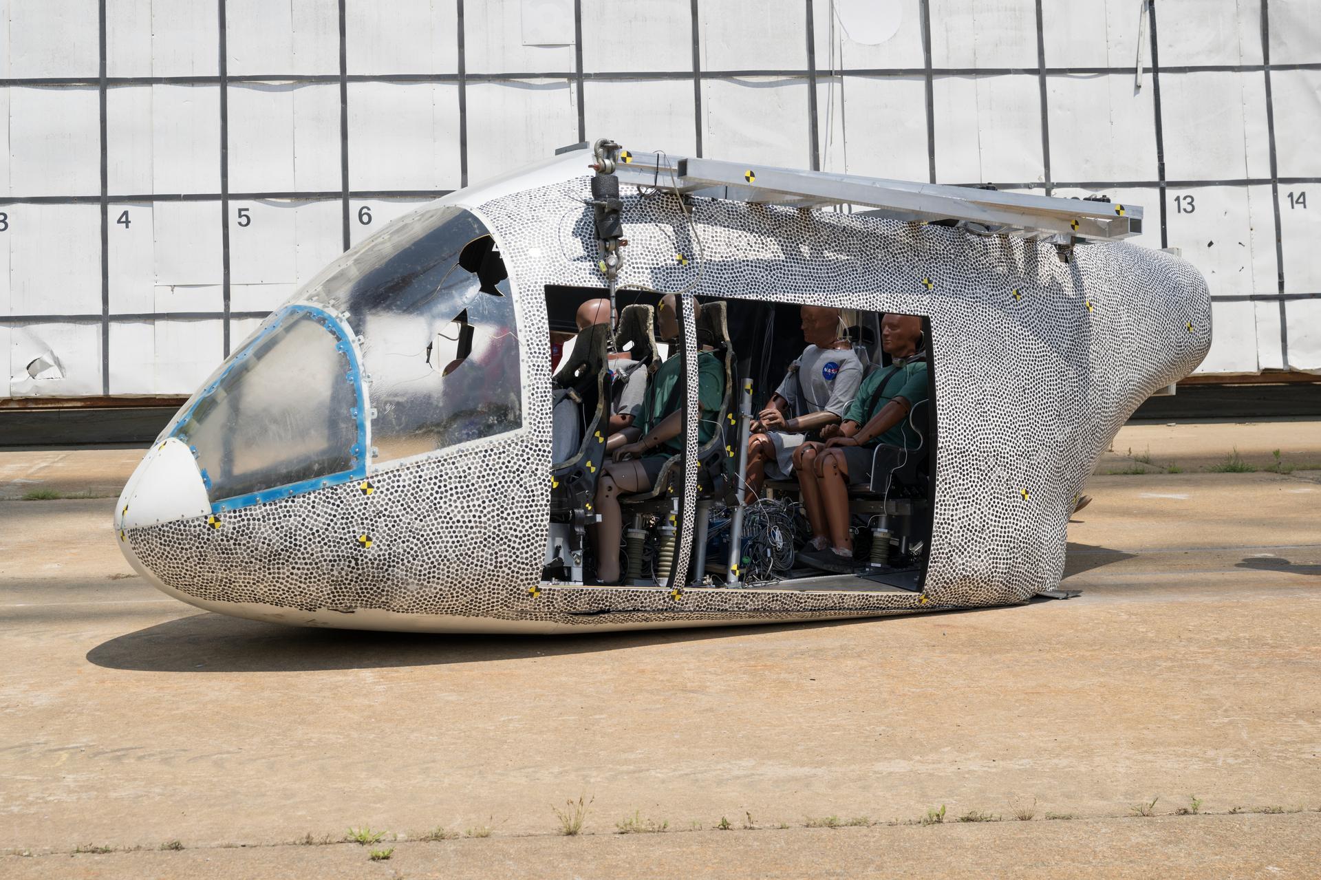 A white aircraft body with small black dots on it sits on the ground after a drop test. The aircraft is damaged on the bottom and there is a hole in the side window. There are several test dummies inside of the aircraft. A steel bar is shown that runs along the top of the aircraft. Behind the aircraft is a large white board to measure the height, which includes many black squares with small numbers inside some of the bottom squares.