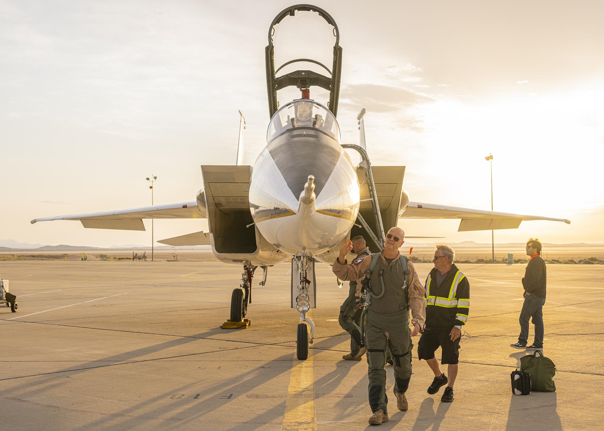 NASA test pilot Nils Larson walks around an F-15B aircraft at NASA’s Armstrong Flight Research Center in Edwards, California. He wears a green G-suit over his tan flight suit and looks up at the nose of the aircraft. The F-15 canopy is open, and support crew stand near the aircraft as the pilots walks around. The aircraft sits higher than the pilot, allowing a partial view of the area on the opposite side.