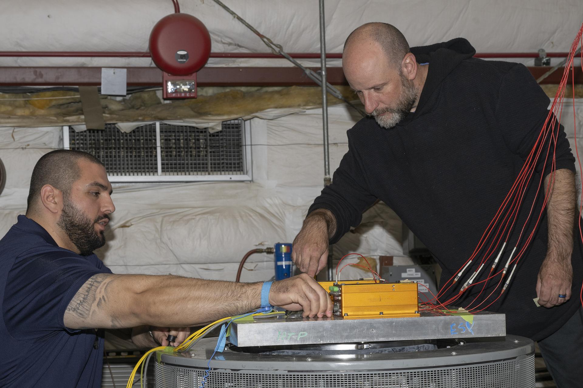 Two men prepare a rectangular system on a rectangular metal surface, which is connected to a cylinder-shaped machine that will severely shake it. Sets of wires are attached to both ends of the rectangular system.