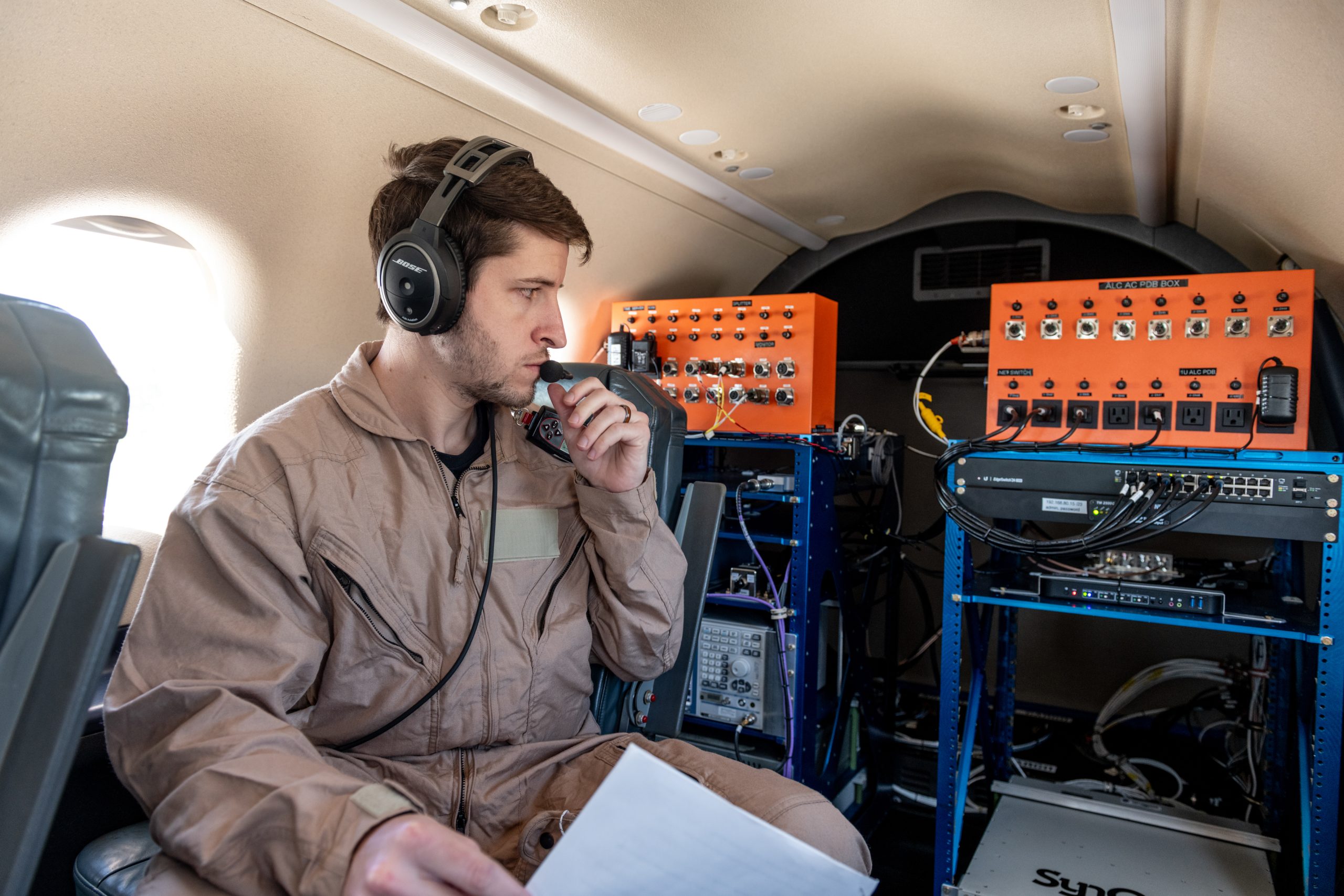 A man in a tan flight suit with a headset and microphone is sitting in a plane holding a piece of white paper. He is looking at communication equipment in the back of the plane. The equipment is comprised of large orange metal boxes with dials connected by wires to a blue computer drive rack.