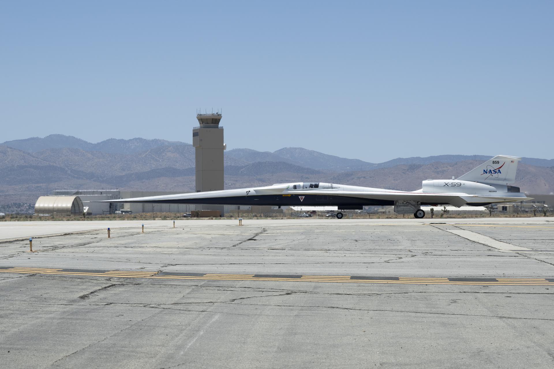 NASA’s X-59 aircraft taxis on the runway at U.S. Air Force Plant 42 in Palmdale, California. The aircraft is shown in profile moving from right to left, with the control tower and desert mountains in the background. In the foreground, the gray concrete runway features a solid yellow line with evenly spaced parallel black stripes inside it, all under a clear blue sky.