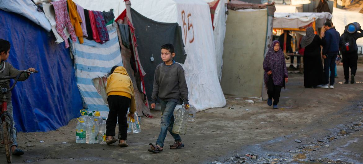 A young boy carries a water bottle through an area where people are living in tents. A young boy carries a water bottle through an area where people are living in tents.