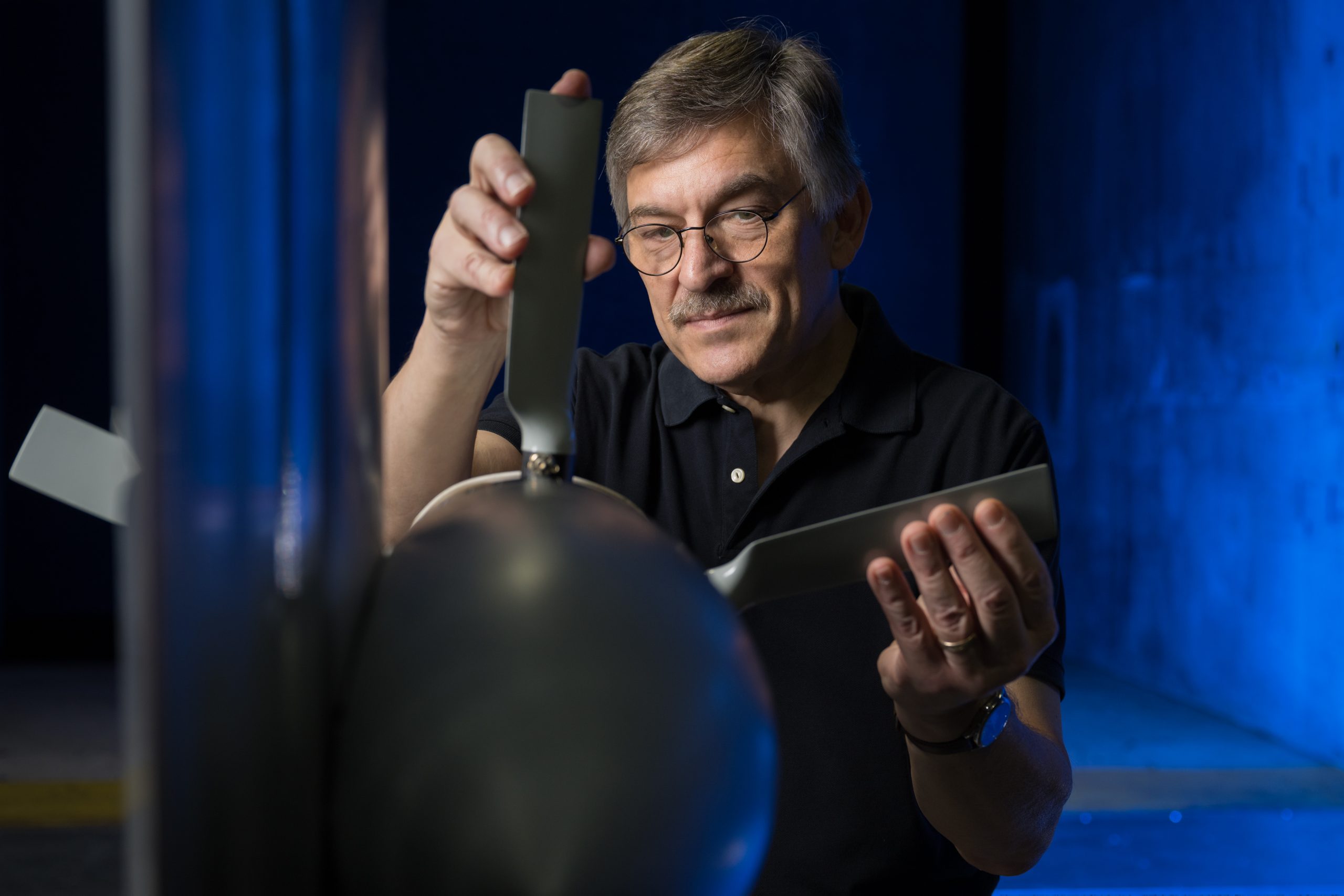 A man is adjusting a propeller blade attached to a test model, with a blue-lit background.