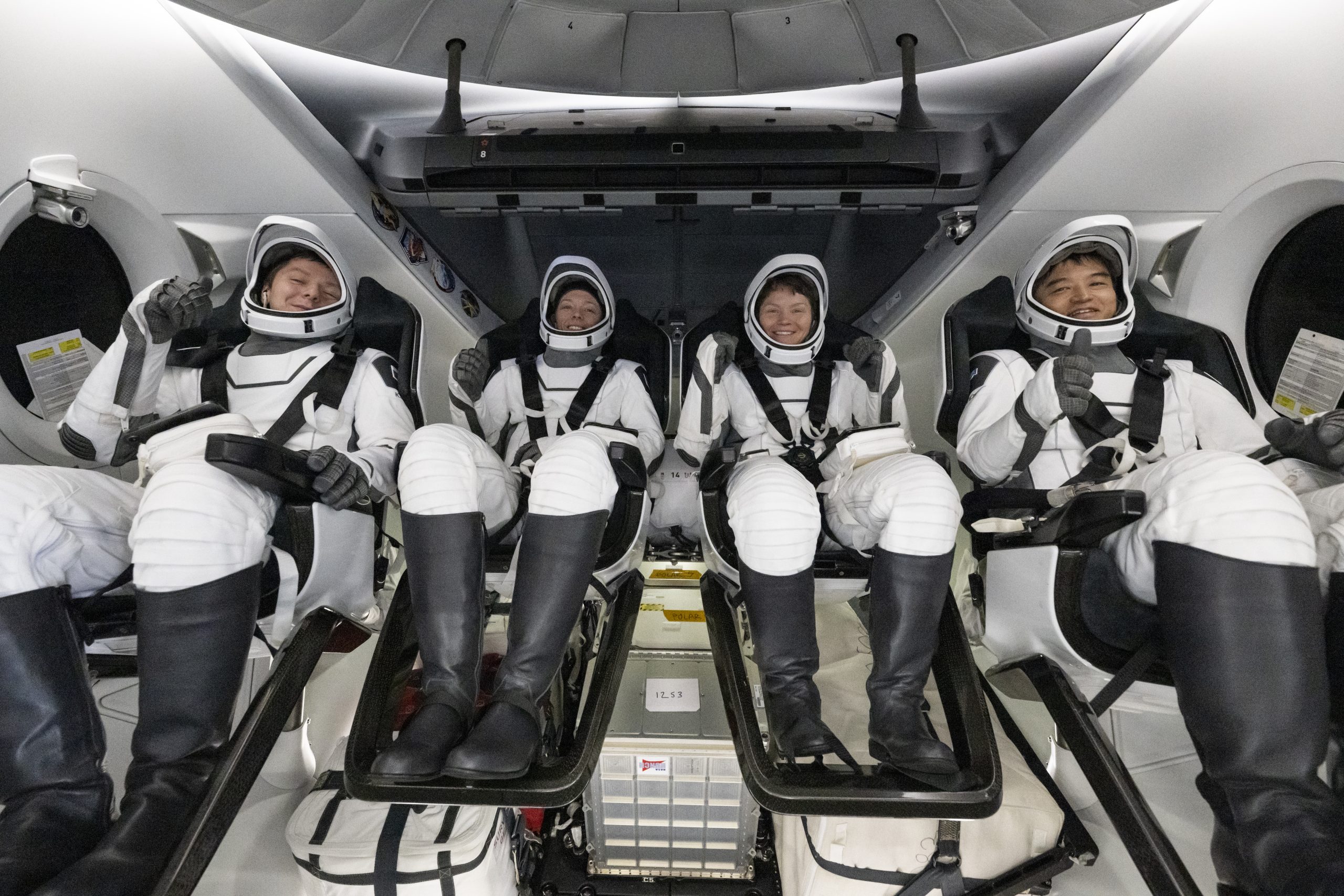 Four people in white suits and helmets with black boots sit in chairs inside a spacecraft that is also white. They are all smiling at the camera.