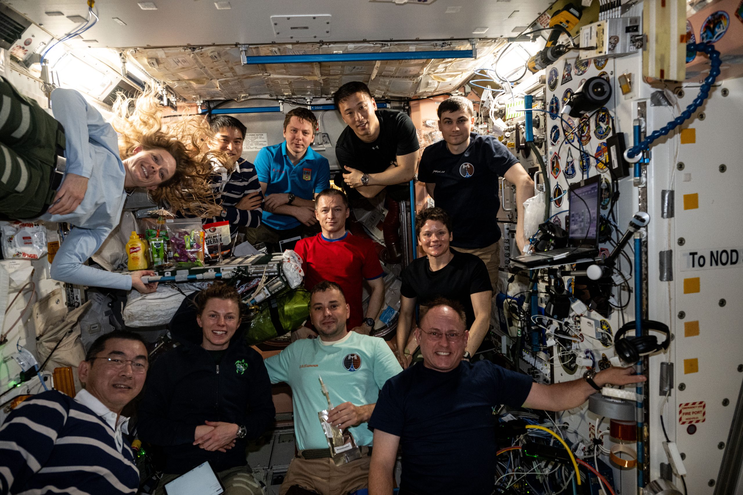 Eleven people pose for a picture in the International Space Station. The crew is composed of women and men from different missions and different countries. The portion of the space station that they're in is white with wires on many of the surfaces.