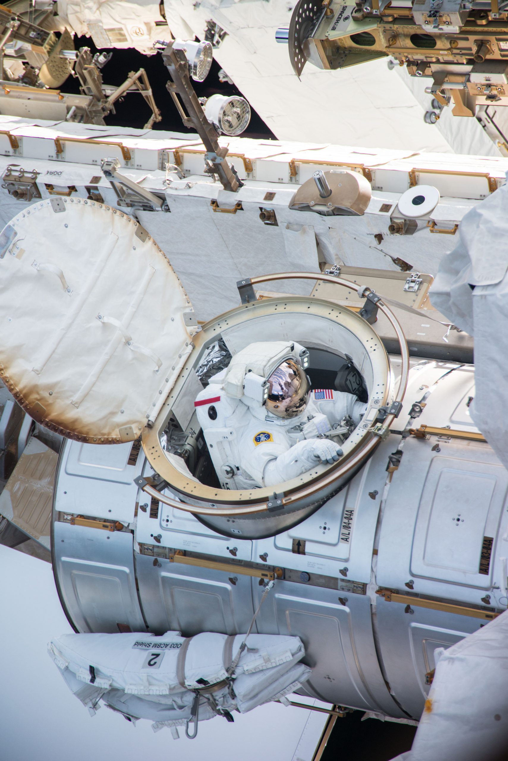 An astronaut in a white spacesuit comes out of a hatch on the International Space Station. We can see parts of the space station in the photo.