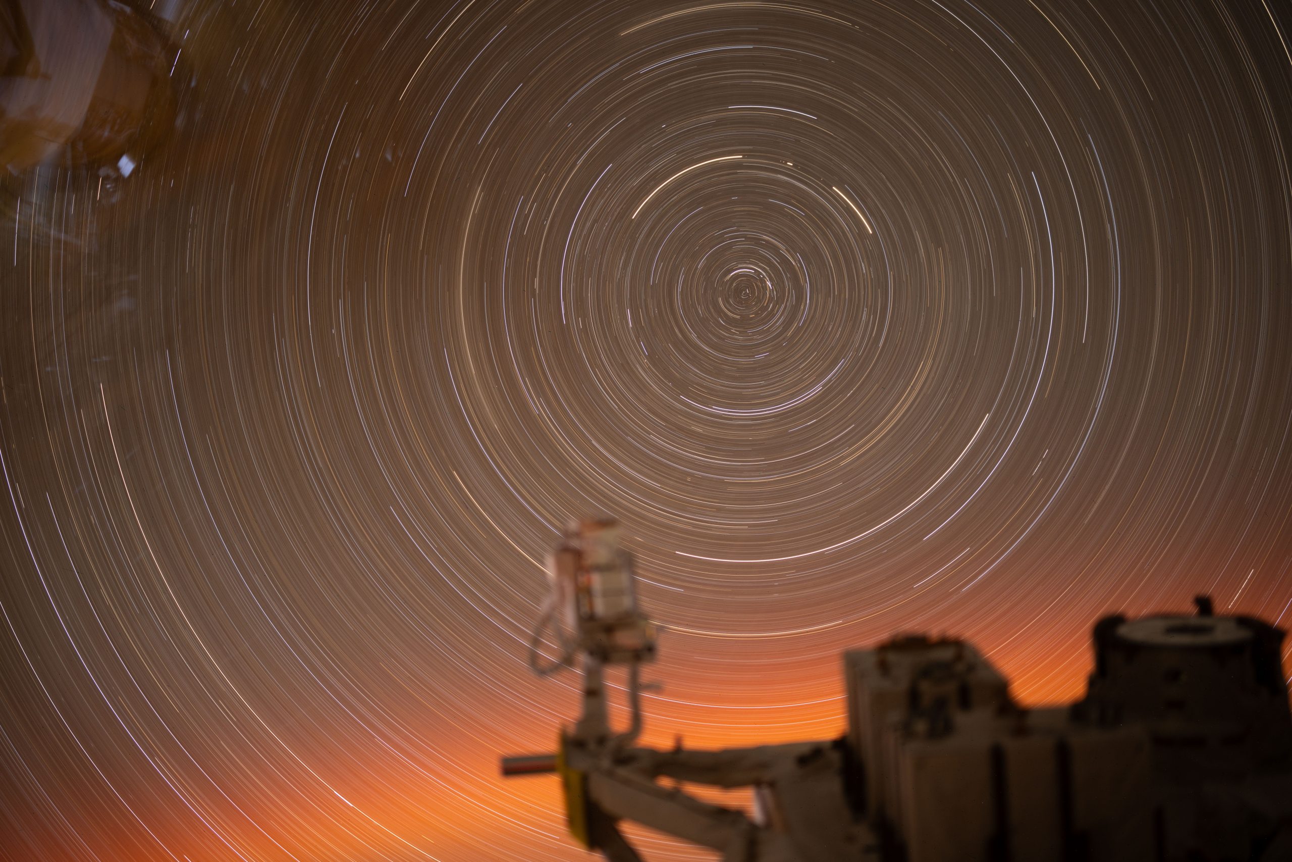 Stars photographed over 31 minutes make a concentric pattern of white circles. At the bottom edge of the photo, there is a soft orange glow. A portion of the International Space Station can be seen in the foreground. This photo was taken through a window on the space station.