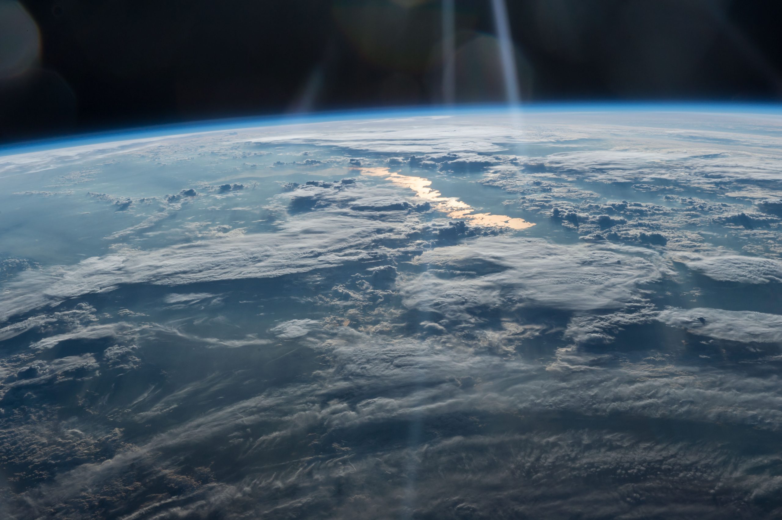 A view of northwestern China (bottom) and eastern Kazakhstan. A body of water in the distance glows gold; this is Lake Balkhash, one of the largest lakes in the world. The rest of Earth that is visible is mostly covered with flat, white clouds. At top, Earth's atmosphere can be seen, as well as the darkness of space.