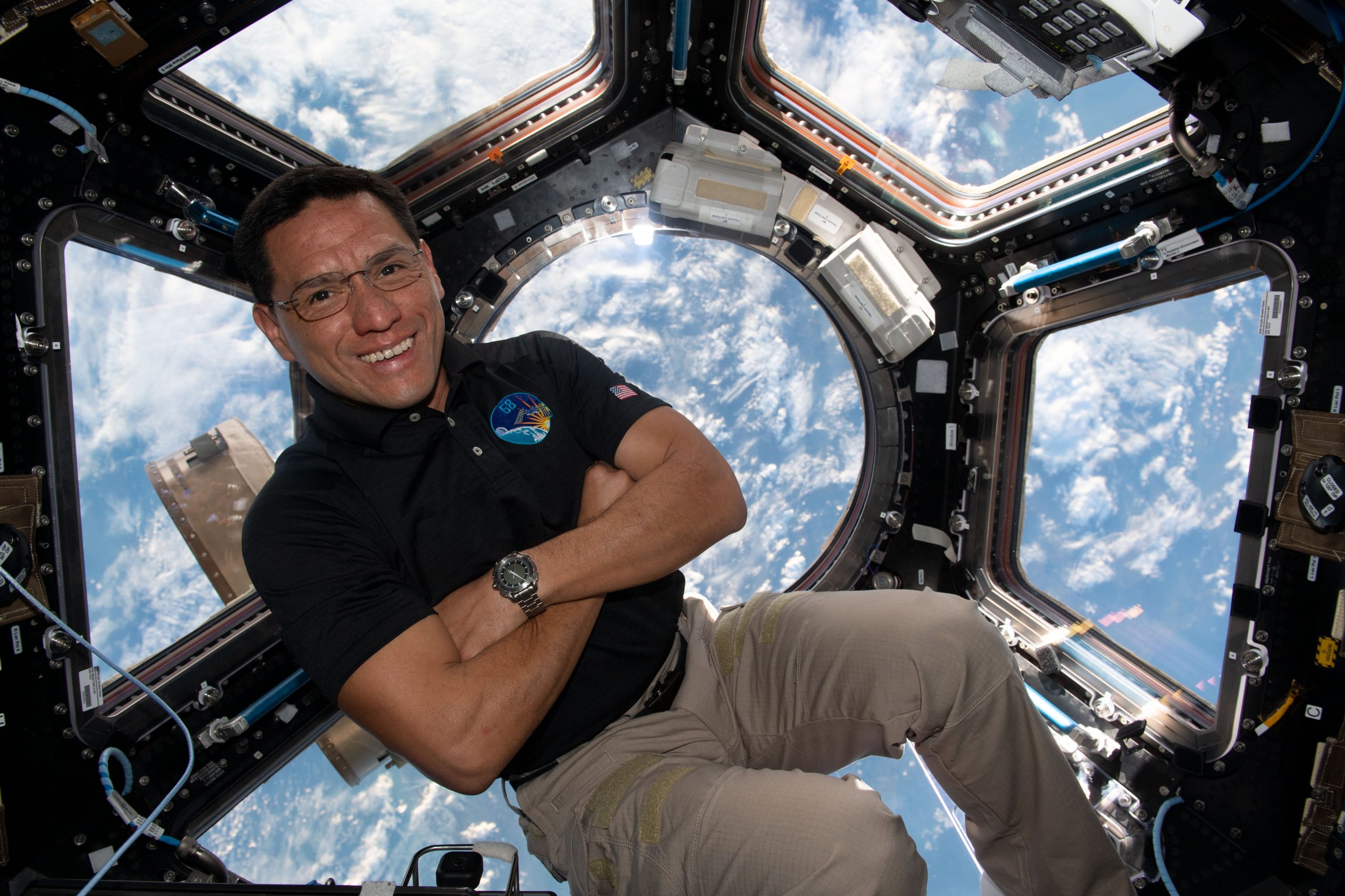 A man in a dark polo shirt smiles at the camera with his arms crossed. He is in the cupola of the International Space Station, an area with multiple windows through which Earth and space can be seen. Earth's clouds can be seen through the windows behind him.