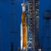 A large orange NASA rocket with white booster rockets stands vertically on a launch pad, lit by spotlights, with the full Moon and night sky behind it.