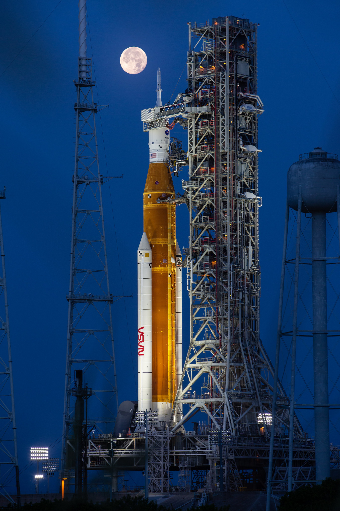 A large orange NASA rocket with white booster rockets stands vertically on a launch pad, lit by spotlights, with the full Moon and night sky behind it.