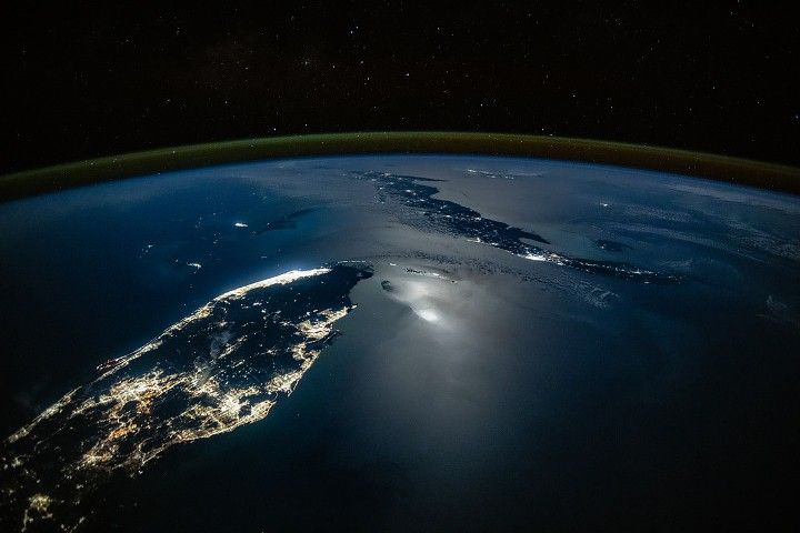 An oblique nighttime photo shows the Florida Peninsula on the left and Cuba on the middle-right, with clusters of city lights illuminating both landmasses. Moonglint shimmers across part of the ocean surface, while green airglow and the darkness of space stretch across the top of the image.