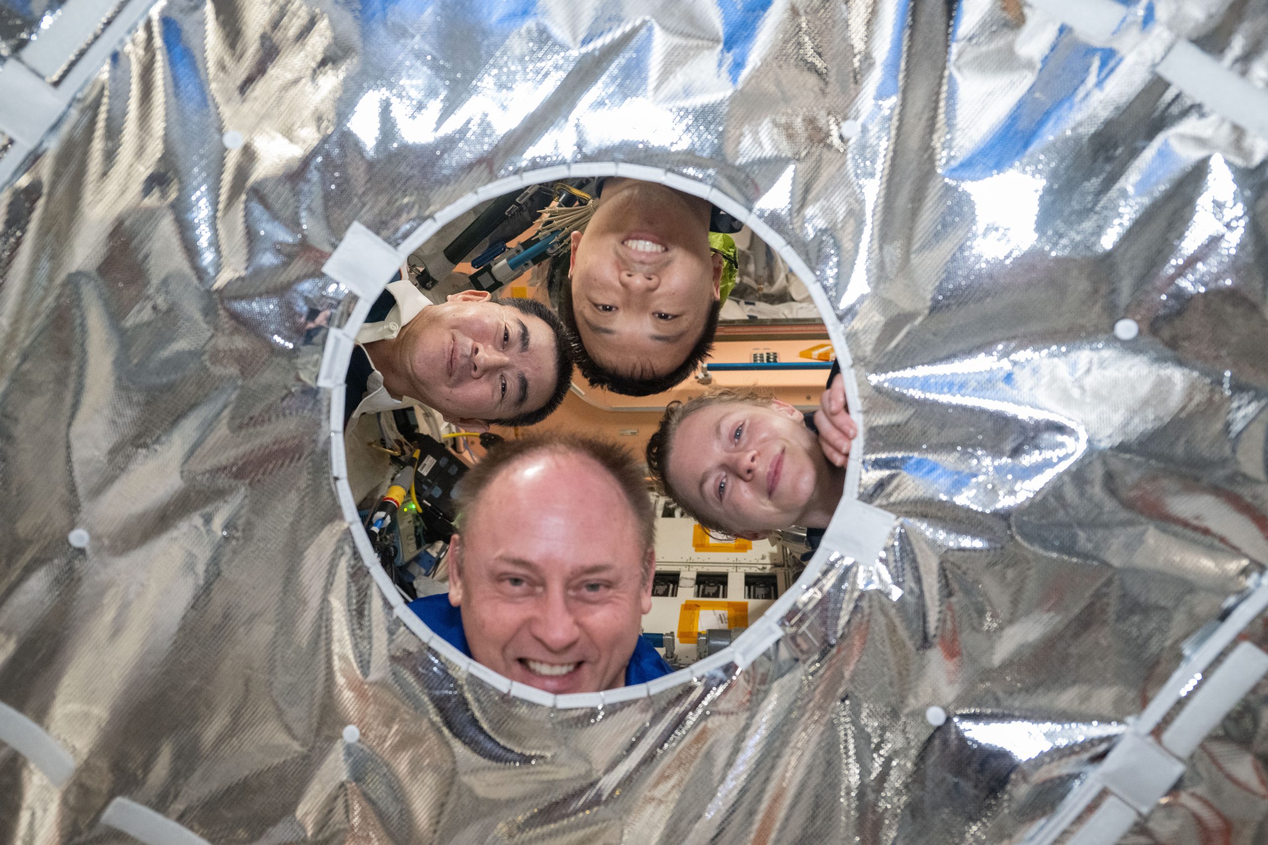 Four people look through a circular hole in a silver metallic fabric. Clockwise from left are JAXA (Japan Aerospace Exploration Agency) astronaut Kimiya Yui and NASA astronauts Jonny Kim, Zena Cardman, and Mike Fincke.