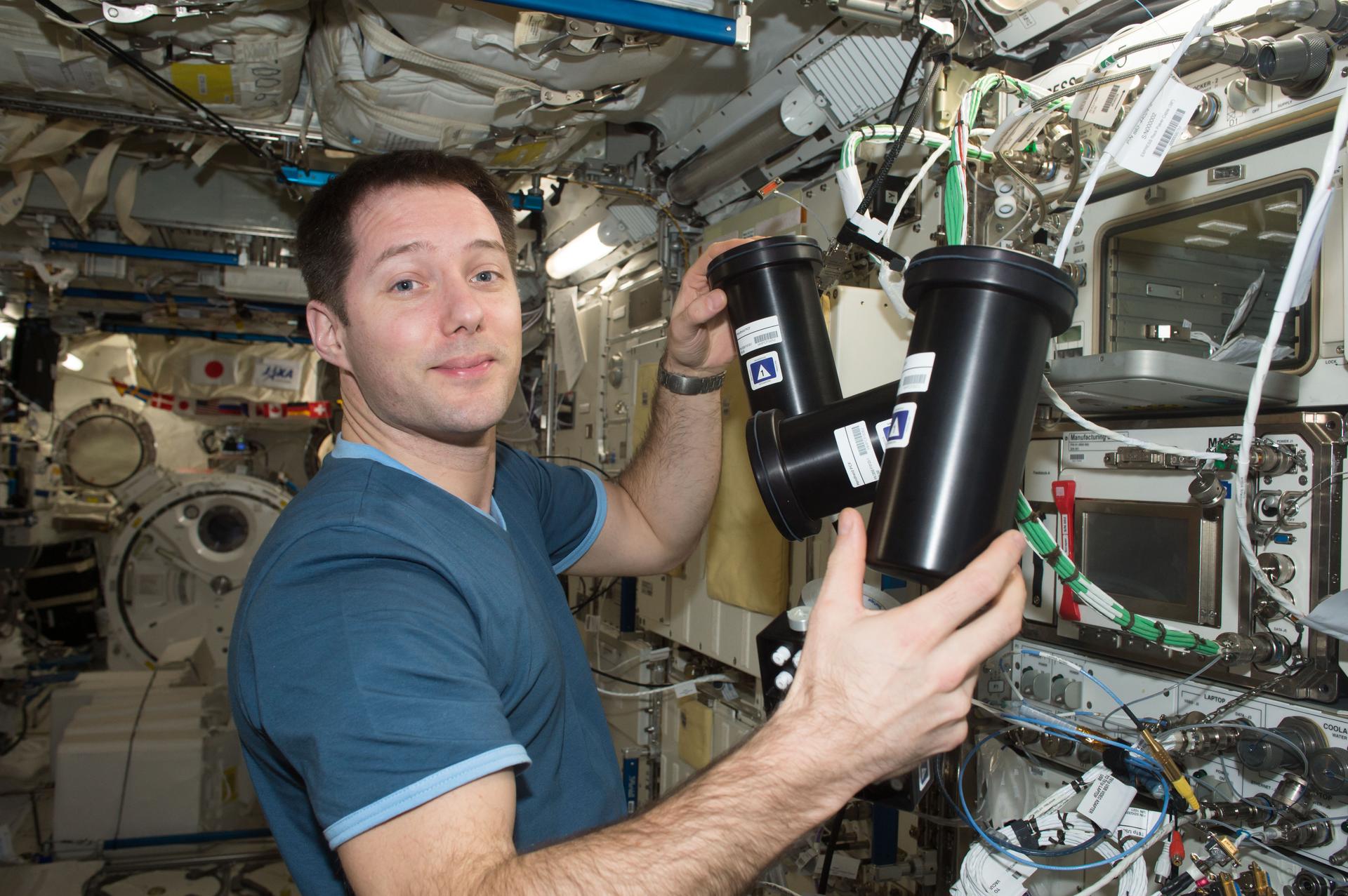 European Space Agency astronaut Thomas Pesquet works inside the International Space Station, . He is holding two cylindrical black experiment containers to deactivate and stow the Protein Crystal Growth-5 hardware. The background is filled withIn the background, cables, silver knobs, instruments, and research equipment are visible.