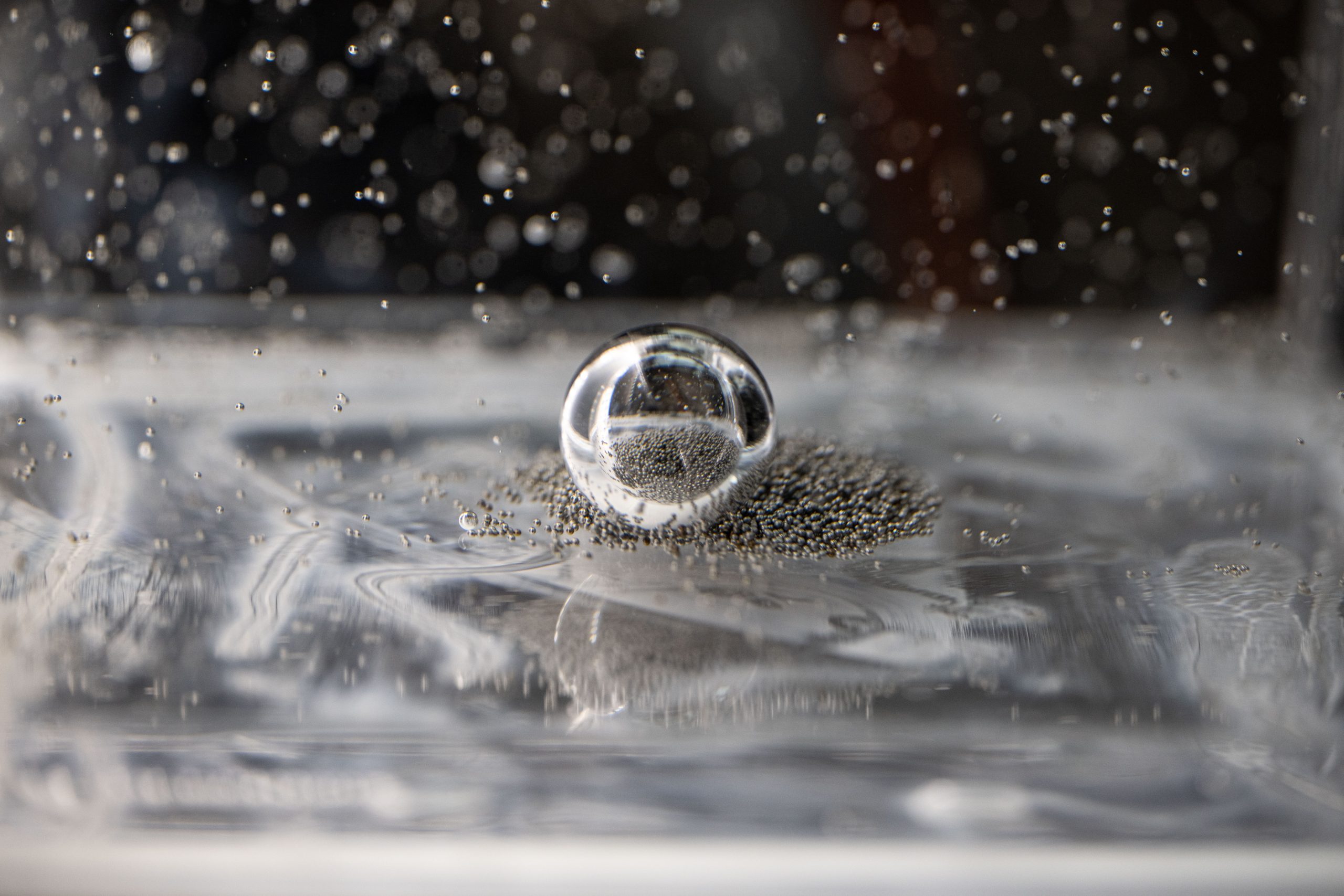 Tiny silver ball bearings crowd around a larger bearing in a clear, viscous fluid. Some of the smaller ball bearings are suspended in the fluid.