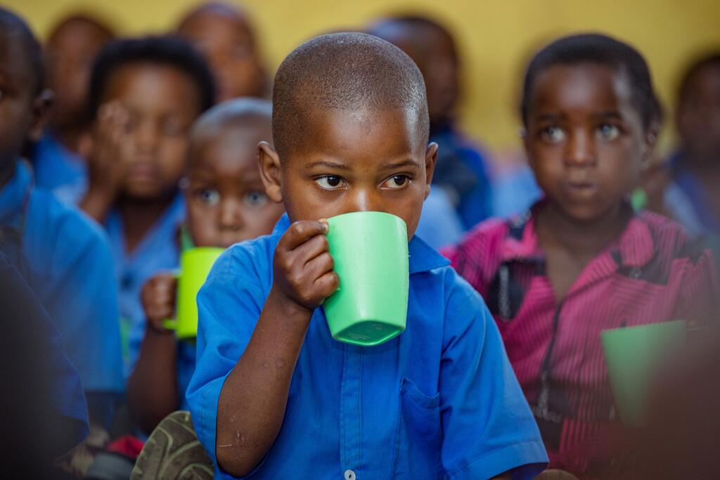 A young child in a blue shirt drinks from a green cup in a classroom setting, surrounded by other children. This image captures a moment from a UNICEF-supported early childhood development initiative in Burera District, Rwanda.