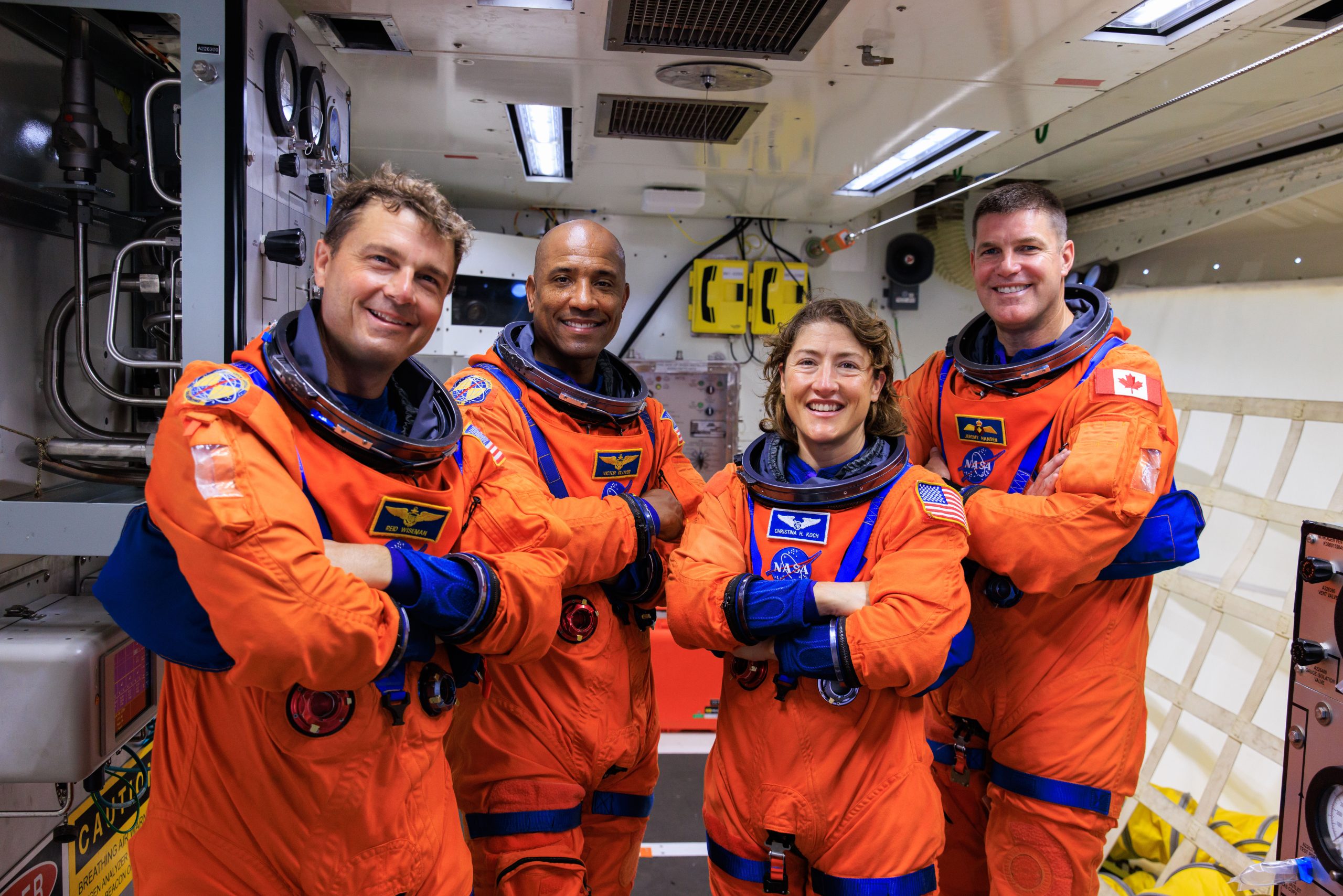 Four astronauts wearing an orange and blue spacesuit pose with their arms crossed inside of a facility.