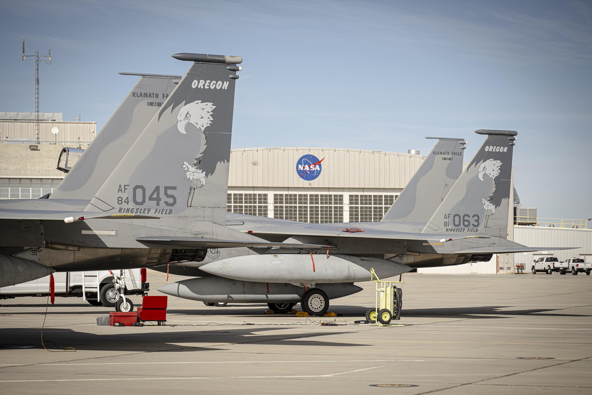 Two F-15 aircraft sit side by side on the ramp at NASA’s Armstrong Flight Research Center, displaying tail numbers 045 and 063 and tail markings that read “Oregon” above an eagle graphic. A NASA hangar with the agency’s logo is visible in the background