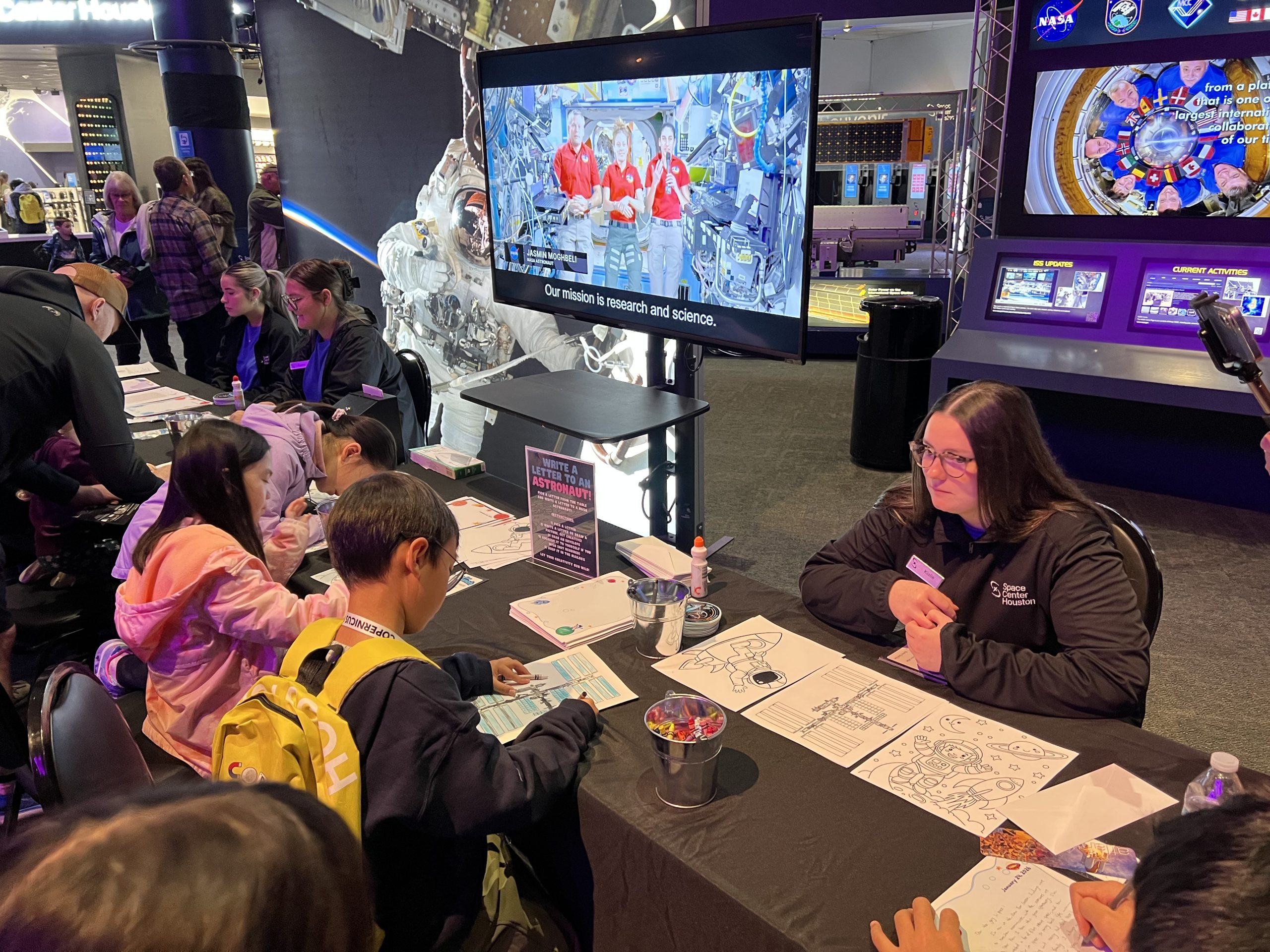 Children participate in a hands-on space activity at Space Center Houston, placing stickers on a poster at an interactive table while visitor center team members provide guidance.