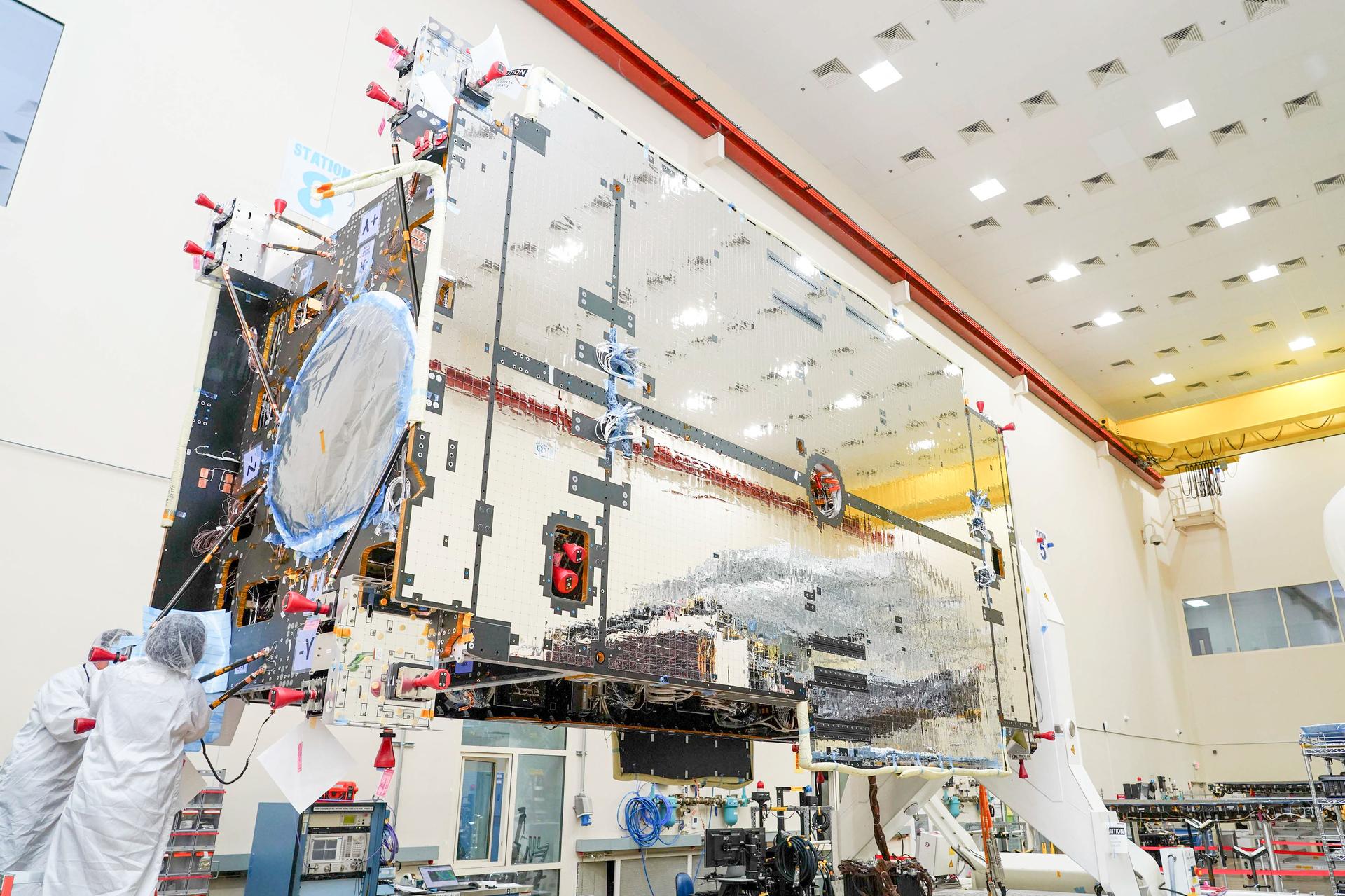 The primary structure of Gateway’s Power and Propulsion Element stands inside a high-bay cleanroom at Lanteris Space Systems in Palo Alto, California. The large rectangular structure is covered in reflective silver-colored panels. Two technicians in white cleanroom suits work near the base of the structure.