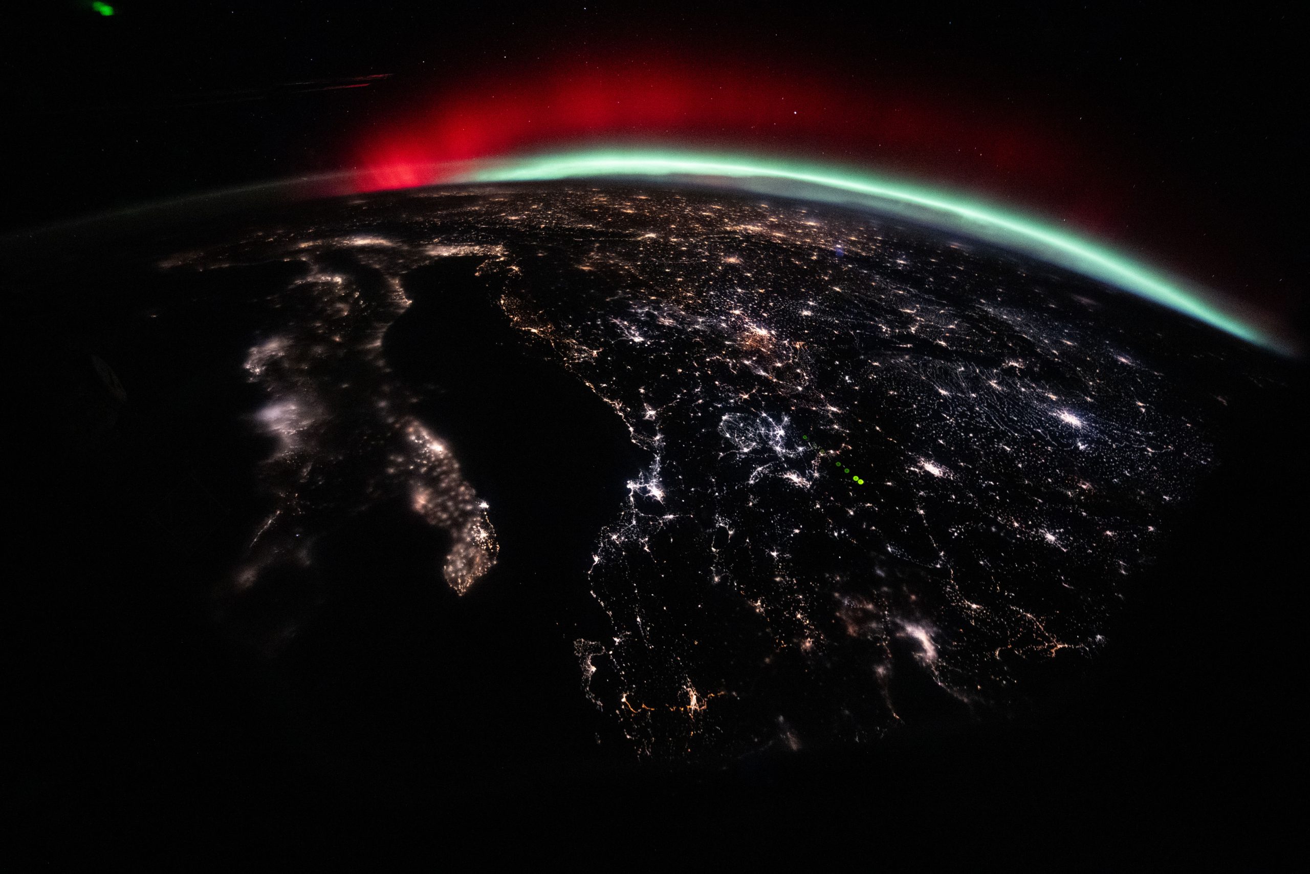 A green and red aurora streams across Earth’s horizon above the city lights of Europe.