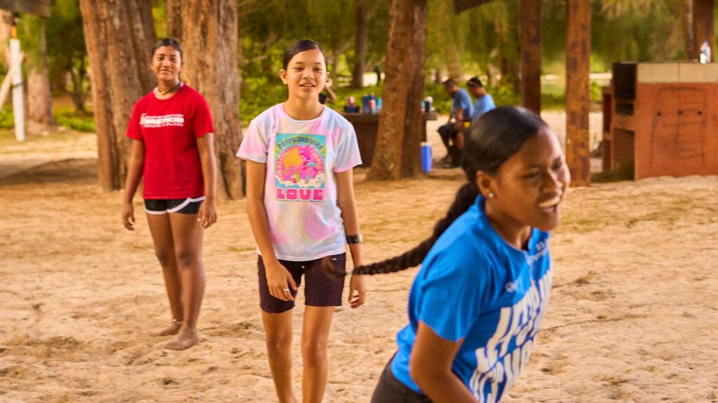 Three young girls in colorful t-shirts play on a sandy beach surrounded by trees in Palau, showcasing community resilience through disaster preparedness.