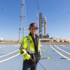 Image shows Jesse Berdis standing standing at the pad of Launch Complex 39B at NASA's Kennedy Space Center in Florida. Behind him are clear blue skies and NASA's SLS (Space Launch System) rocket and Orion spacecraft for the Artemis II mission. Photo credit: Kim Shiflett