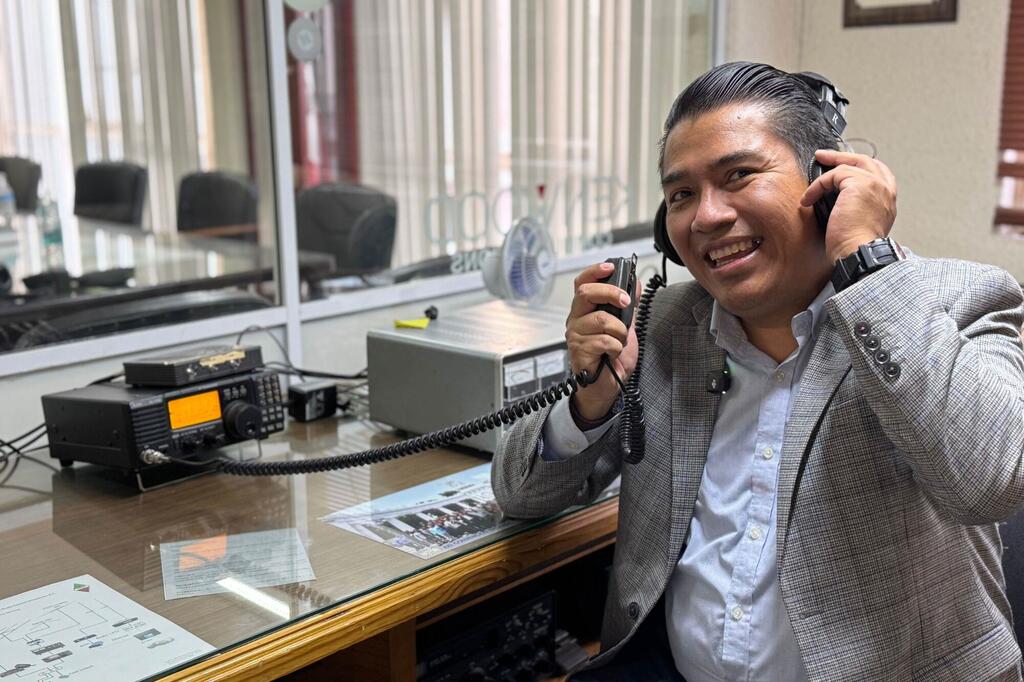 A smiling male news anchor wearing headphones and holding a radio microphone in a studio setting, with radio equipment visible on the desk.