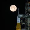 A golden full moon is seen shining over NASA’s SLS (Space Launch System) and Orion spacecraft, atop the mobile launcher in the early hours of February 1, 2026. The SLS core stage is a distinctive deep orange, while Orion is white and sits on top of the core stage.