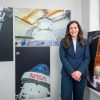 A professionally dressed woman stands in a gallery style room surrounded by large NASA photographs showing the Orion spacecraft in space, engineers working on a module, a close-up of the Orion spacecraft with the Moon behind it, and the SLS (Space Launch System) rocket illuminated on the launch pad.