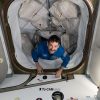 A man with dark hair and a mustache looks up at the camera, smiling. His hands are on the bottom edge of the hatch entrance as he makes his way inside the space station. The path he is coming through has many square cargo bags strapped to each side.