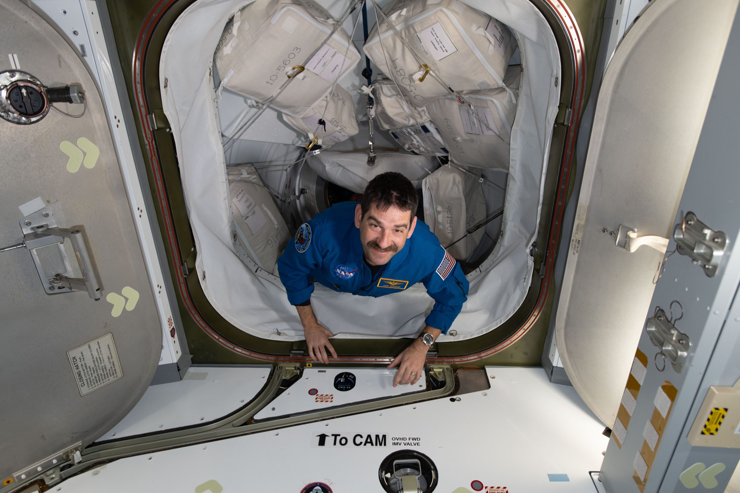 A man with dark hair and a mustache looks up at the camera, smiling. His hands are on the bottom edge of the hatch entrance as he makes his way inside the space station. The path he is coming through has many square cargo bags strapped to each side.