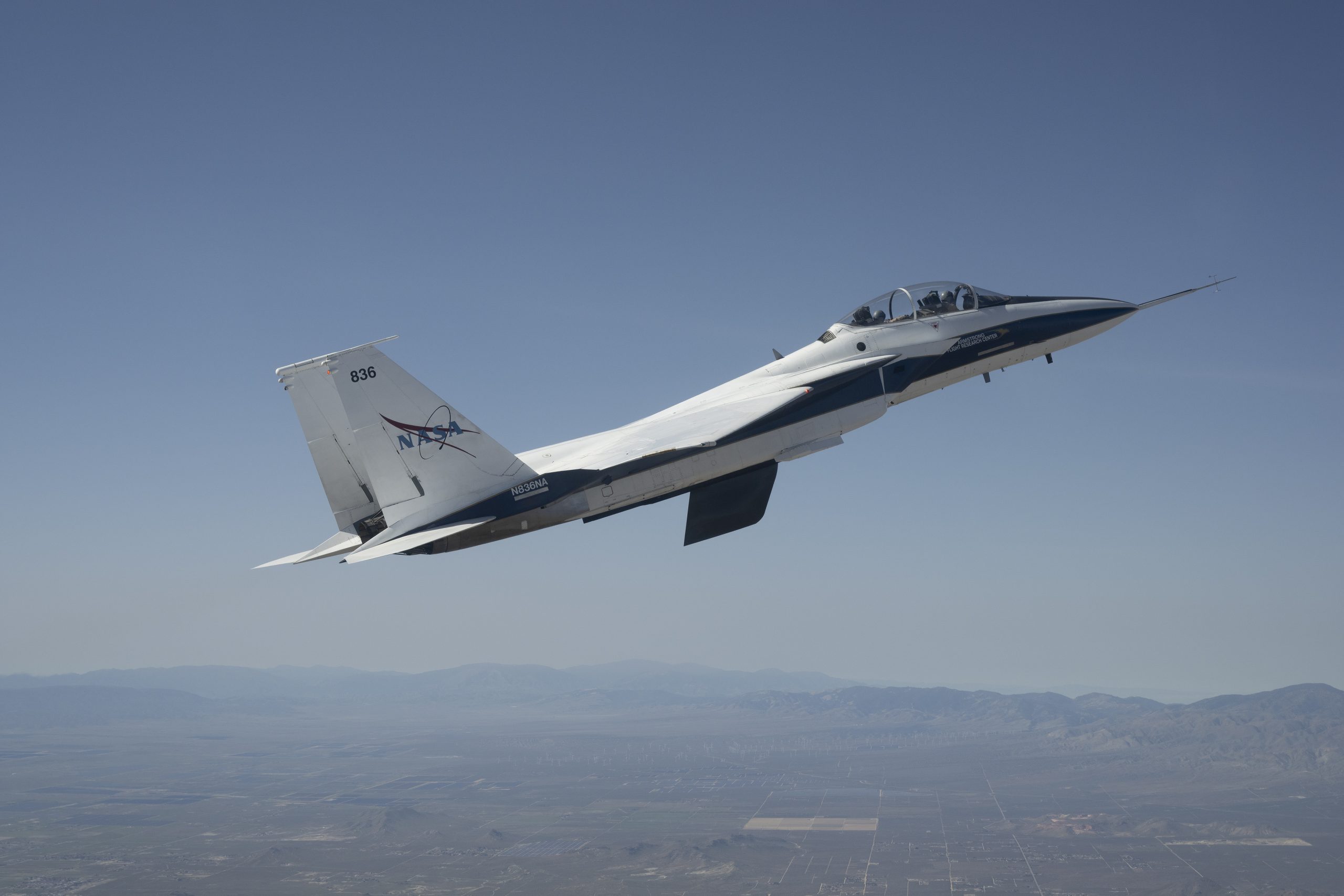 A white and blue NASA F-15 research jet climbs to altitude with an approximately 3-foot experimental wing design mounted beneath its fuselage. Viewed in profile against a blue sky with mountains in the distance, the test article resembles a ventral fin below the aircraft.