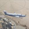 NASA’s freshly painted Pilatus PC-12 flies over NASA’s Armstrong Flight Research Center in Edwards, California. The parking lots and center buildings dot the landscape below. The compass rose in the upper part of the photo shaped like the sun hovers over the aircraft, emphasizing the gleaming white plane with a blue stripe and blue N606NA number across the fuselage and NASA red worm logo on the tail. A sensor, with a black-tip juts out from below the wing.