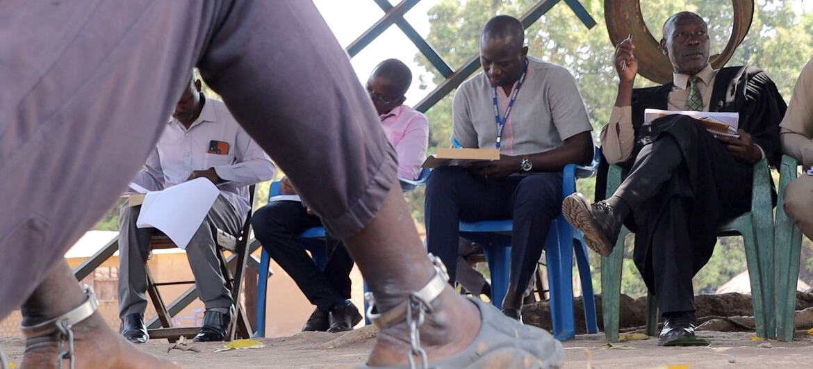 A close-up of a person's feet in chains, with a mobile court in the background in South Sudan, highlighting the justice system's efforts to address serious criminal cases.