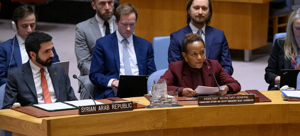 A woman in a maroon blazer speaking at a conference table with a nameplate reading 'ASSISTANT SECRETARY-GENERAL HUMANITARIAN AFFAIRS AND DEPUTY EMERGENCY RELIEF COORDINATOR'. Behind her, a man sits at a table with a nameplate for the 'SYRIAN ARAB REPUBLIC'.