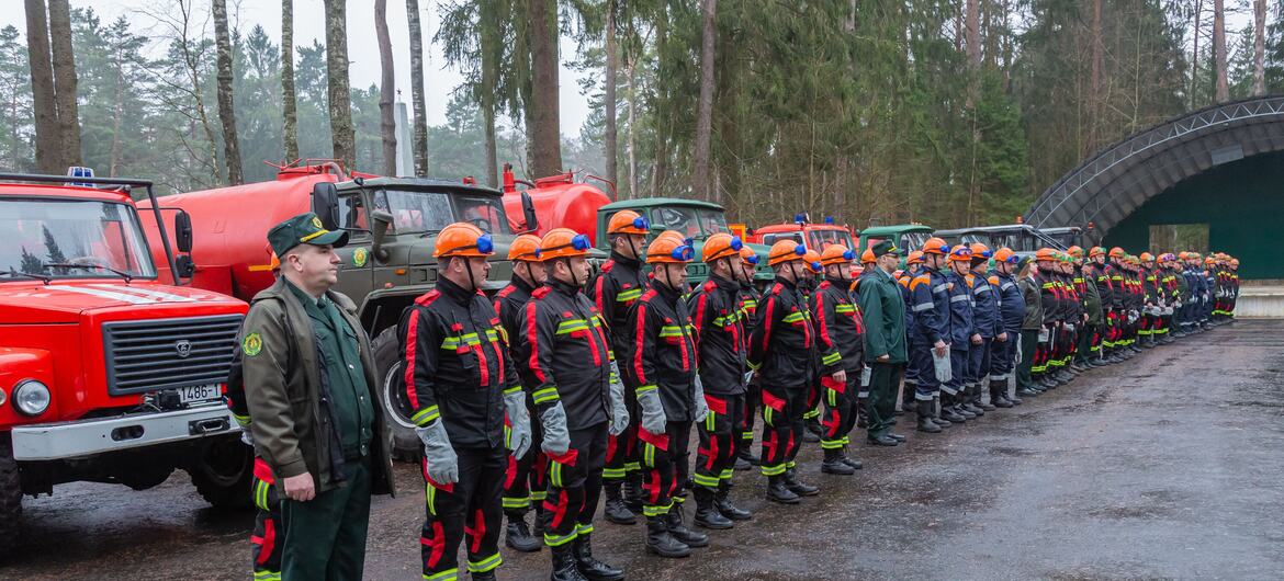 A line of Belarusian firefighters in uniform and orange helmets standing in front of fire trucks in a forest.