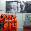 Four neon orange spacesuits hang on a rack decorated with NASA patches. Three of the suits have American flag patches on their left shoulder, while the last has a Canadian flag patch on its left shoulder. The room the suits are hanging in has a large black and white photo on the wall, in which astronauts are in their spacesuits and helmets while two people in white lab coats look on.