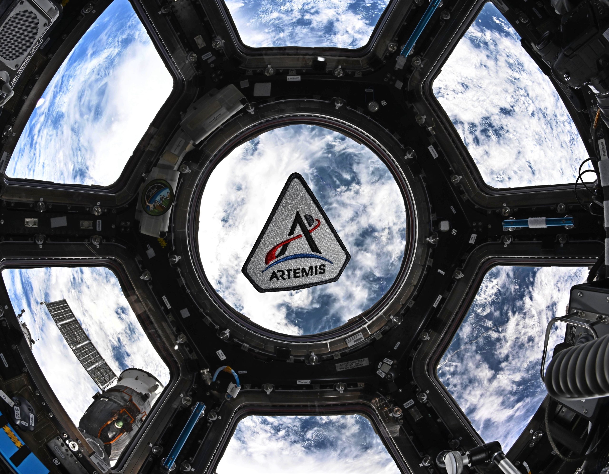 The Artemis program patch in the space station's cupola. The patch is framed by the center circular window in the space. Earth can be seen behind the patch. The Artemis patch has five sides. It has a white background with a black border. There's a black "A," for Artemis. The crossbar of the A is made by a curving red trajectory line that crosses from left to right. That line continues beyond the "A" and beyond a small gray circle representing the Moon. Below the "A" is a blue crescent representing Earth. At the very bottom is the word "Artemis."