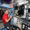 NASA astronaut Chris Williams is seated inside the Destiny laboratory module aboard the International Space Station. He is wearing a red shirt and black shorts with three white stripes underneath the right pocket. Chris is holding a device with a cord in his left hand and there are blank computer screens in front of him. He is smiling brightly and looking directly at the camera.