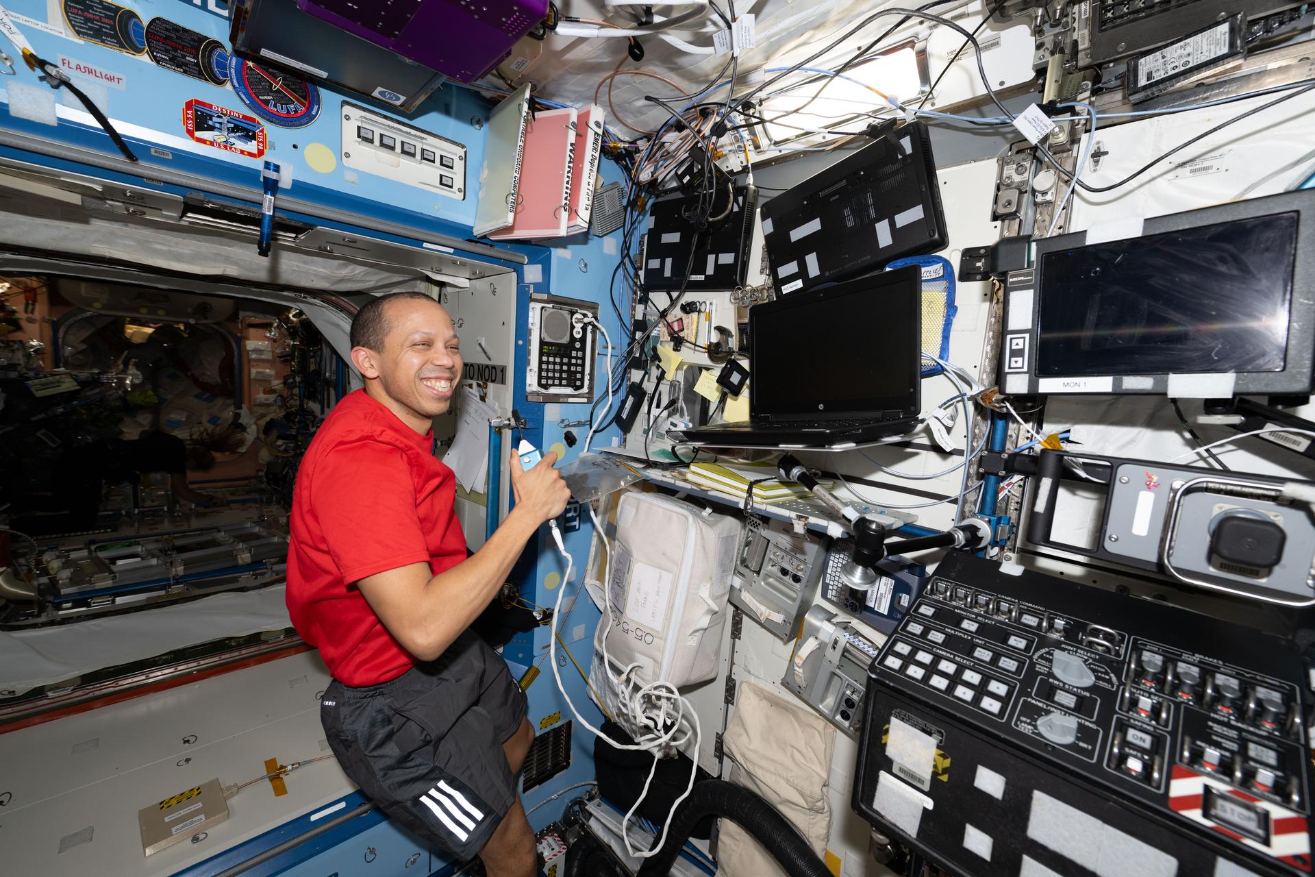 NASA astronaut Chris Williams is seated inside the Destiny laboratory module aboard the International Space Station. He is wearing a red shirt and black shorts with three white stripes underneath the right pocket. Chris is holding a device with a cord in his left hand and there are blank computer screens in front of him. He is smiling brightly and looking directly at the camera.