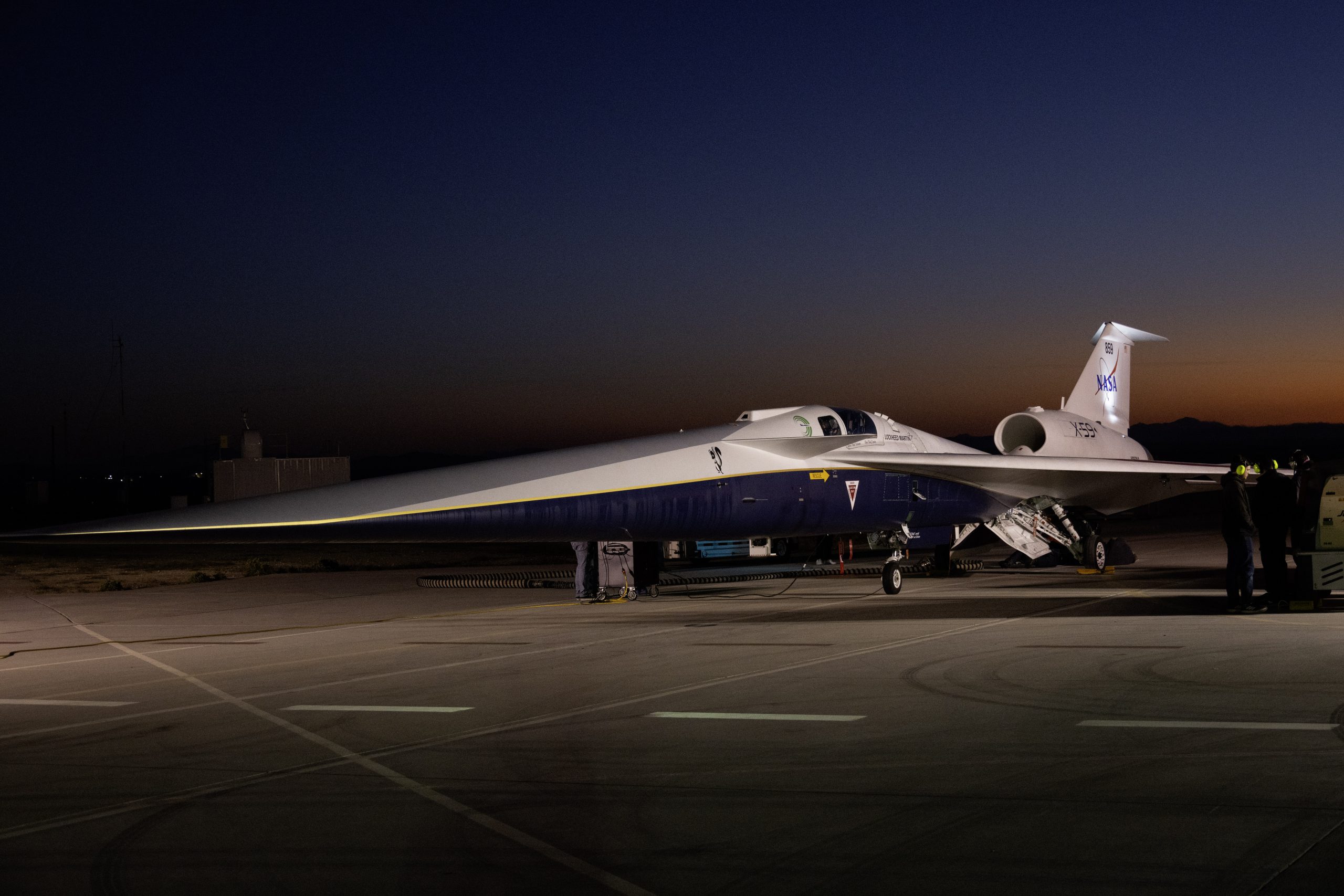 An aircraft resting on a section of runway as seen from the side. The X-59 has a long, thin nose that accounts for nearly a third of its length, along with sleek wings and an engine mounted above its body, just below its tail. The early morning sky is dark in the background with the sunrise just starting to emerge.