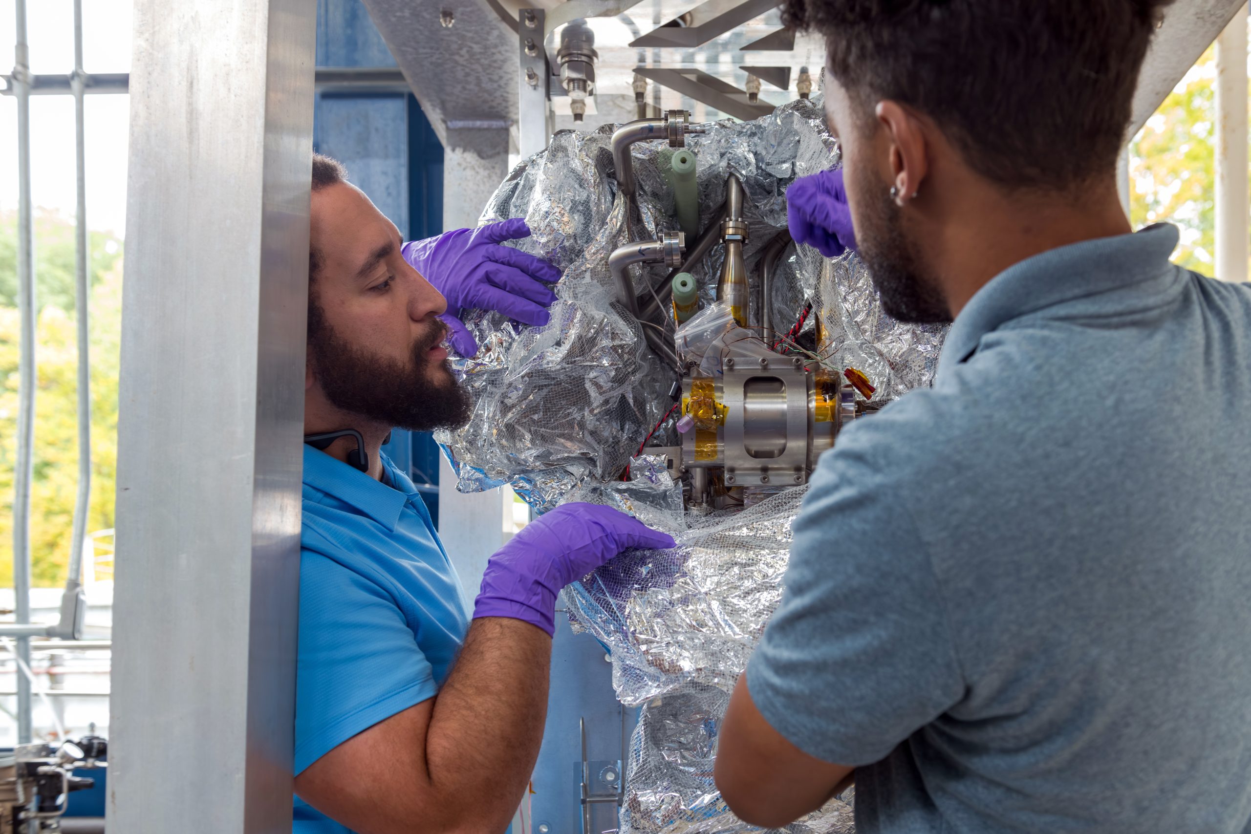 Two engineers wearing purple gloves peel away wrapping around metal and yellow hardware. The hardware is mounted inside a steel metal frame. This technology could help NASA fuel future missions,