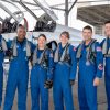 From left to right, NASA astronauts Andre Douglas, Victor Glover, and Christina Koch, CSA (Canadian Space Agency) astronauts Jenni Gibbons, NASA astronaut Reid Wiseman, and CSA astronaut Jeremy Hansen pose in front of an airplane. They are all wearing blue jumpsuits with patches and gray harnesses.