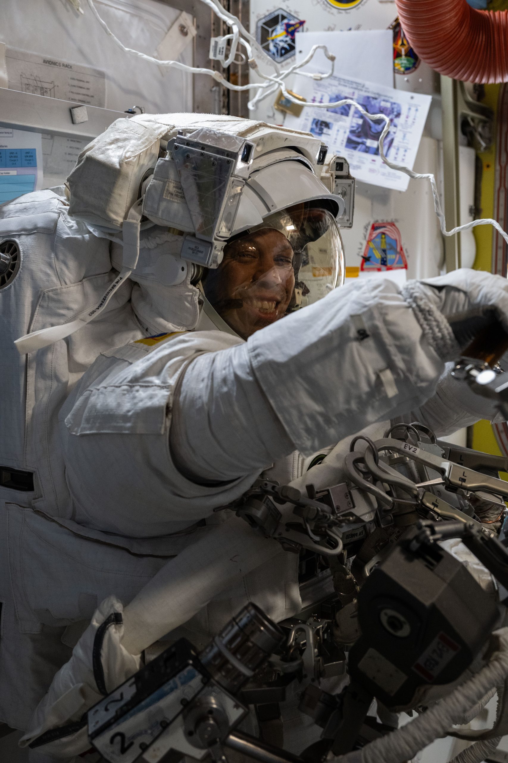 Chris Williams, an astronaut, is wearing a white spacesuit and helmet. He is facing the camera, but his body is turned to the right of the image. His arms are extended in front of him.