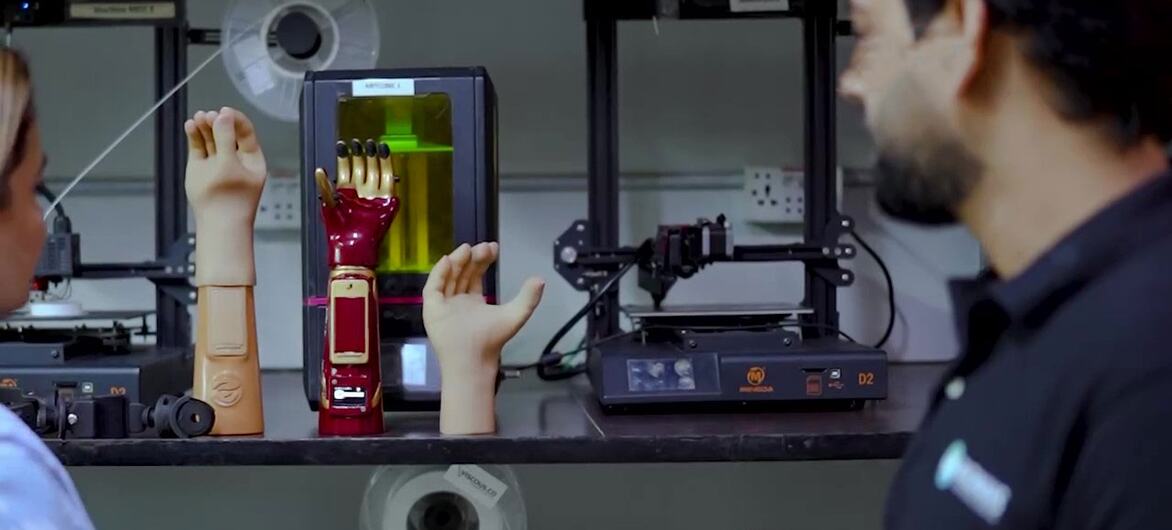 Two individuals observing AI-powered prosthetic limbs displayed on a workbench with 3D printers in the background.